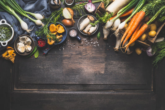An arc of vegetables and spices, including carrots, garlic, spring onions, mushrooms, parsnips, potatoes, fennel, salt, peppercorns, and rosemary, arranged on a dark-stained wooden board.